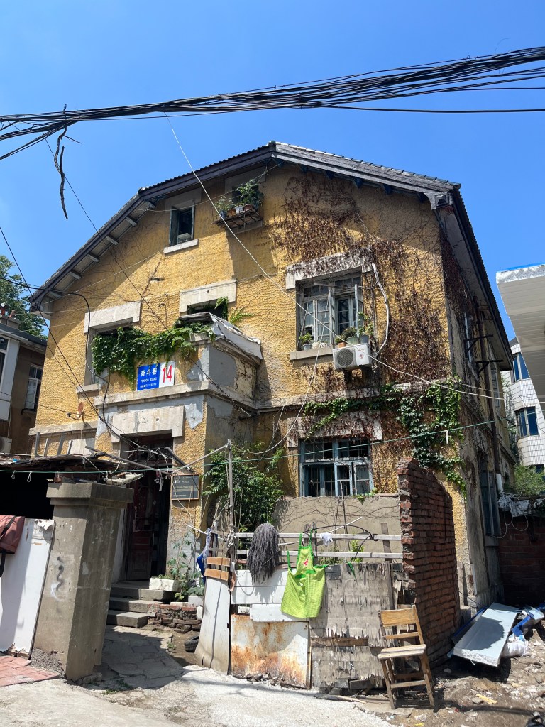 古い黄色い家の外観。窓には植物が絡みつき、周囲には洗濯物が干されている。