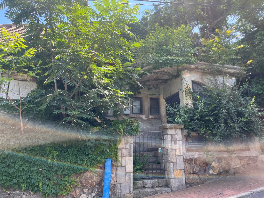 緑に覆われた古い家の外観。石の壁と木のドアが見える。日差しが差し込み、風景が明るく照らされている。
