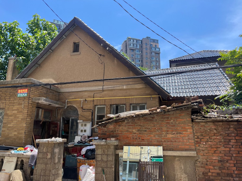古い家屋の外観、青空の下で、周囲にはブロック壁や雑多な物品が置かれている。